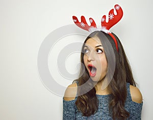 Surprised young woman with reindeer horns on her head looks your pruduct / object with mouth open on gray background. Copy space.