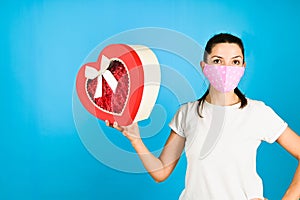 Surprised young woman in white t-shirt and protective mask opening a gift box isolated over blue background. Sales concept