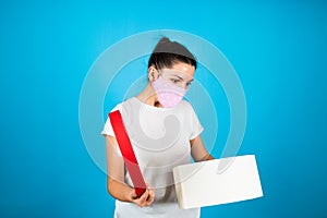 Surprised young woman in white t-shirt and protective mask opening a gift box isolated over blue background. Sales concept