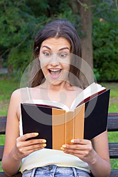Surprised young woman with widely open yeas and mouth is reading a book outdoors