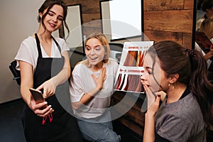 Surprised young women looking at phone while hairdresser holding it in hand. Hairstylist proposing fashion hair coloring