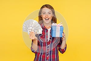 Surprising present, money bonuses. Portrait of amazed excited ginger girl in casual checkered shirt holding gift box and dollars