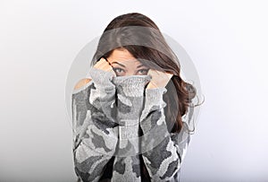 Surprising scared woman with big open eyes hiding her face inside the grey warm winter pullover on blue background