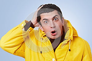 Surprsied desperated handsome man looks with bugged eyes and widely opened mouth, wears yellow anorak with hood, can`t understand