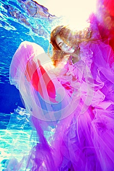 A surreal portrait of a teenage girl underwater in a luxurious multi-colored dress against the background of bright