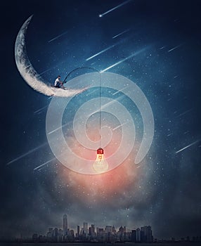 Surreal scene over the night city with a creative boy seated on the crescent moon using a fishing rod with a light bulb as a bait