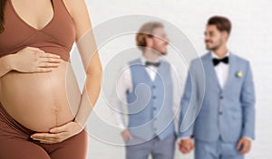 Surrogacy concept. Young pregnant woman and blurred view of happy gay couple on light background