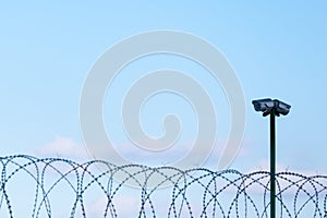 Surveillance cameras over barbed wire against a blue sky. Fenced guarded territory, no access. Area protection and counteraction