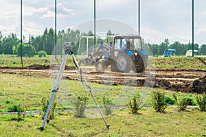 Surveying measuring equipment level theodolite stands on a tripod on a green lawn, in the background a tractor levels the ground