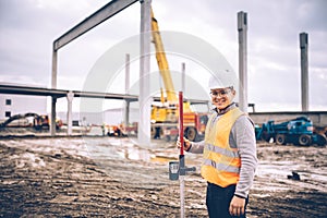 Surveyor engineer smiling with surveying tools and equipment at construction site outdoors, prefabricated cement pillars and beams