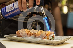 sushi chef preparing maki sushi roll with torch burner, firing direct towards smoke salmon in the kitchen of izakaya