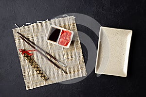 Sushi chopsticks, soy sauce, ginger, bamboo mat and empty plate on black background.