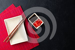 Sushi chopsticks, soy sauce, ginger, red bamboo mat and empty plate on black background.