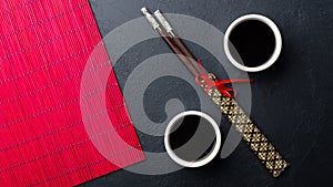 Sushi chopsticks, soy sauce, ginger, red bamboo mat and empty plate on black background.