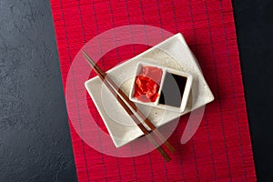 Sushi chopsticks, soy sauce, ginger, red bamboo mat and empty plate on black background.