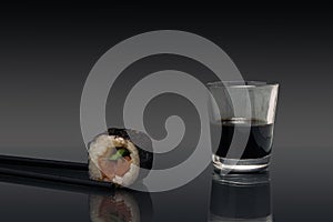 Sushi minimal concept. Chopsticks, one piece of rice, salomon and cucumber roll in nori dried edible seaweed beside soy sos glass