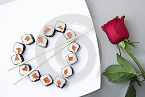 Sushi rolls laid out in the shape of a heart on a white plate. Red rose on a gray background. Creative greetings. The concept of