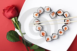 Sushi rolls laid out in the shape of a heart on a white plate. Red rose on a red background. Creative greetings. The concept of