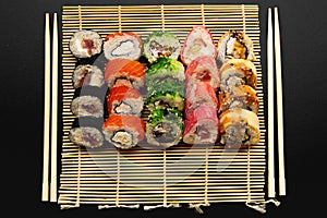 Sushi set with different maki - hosomaki, futomaki and uramaki on bamboo mate with chopsticks on a black background