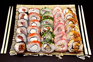 Sushi set with different maki - hosomaki, futomaki and uramaki on bamboo mate with chopsticks on a black background