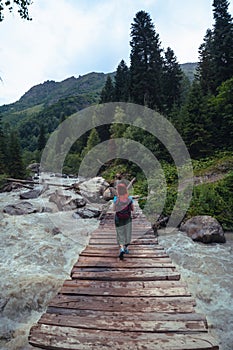 A suspension bridge on hiking trail through green dense forest with a female traveler. Woman tourist standing on wooden bridge