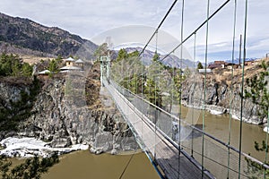 Suspension hanging bridge above water mountain river stream covered with ice snow on the background of rock island, Katun river,