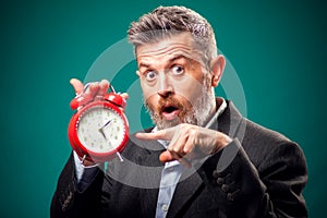 Susprised bearded businessman holding alarm clock and pointing with a finger at it. Time management concept