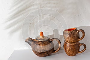 Sustainable hand crafted pottery tea set, teapot and cups standing on white kitchen shelf, floral sunlight shadow on wall, copy