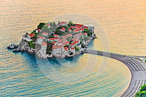 Sveti Stefan small island with red-tiled roofs, green trees and beautiful sandy beach with sunbeds on the Adriatic coast of photo