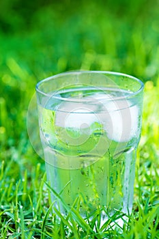 Sweated frosty glass with clear pure cool water with ice cubes on green grass background. Hydration summer refreshment