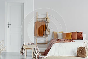 Sweaters, wicker hat and bag on wooden ladder next to king size bed with velvet pillows in white bedroom interior, copy space on