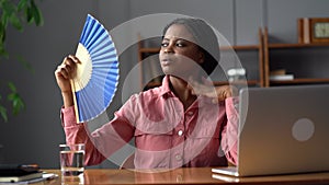 Sweaty african woman worker sit at laptop at hot office try to avoid overheating using paper fan