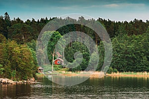 Sweden. Beautiful Red Swedish Wooden Log Cabin House On Rocky Island Coast In Summer. Lake Or River And Forest Landscape