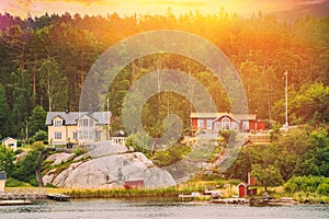 Sweden. Beautiful Red And Yellow Swedish Wooden Log Cabins Houses On Rocky Island Coast In Summer Evening. Lake Or River