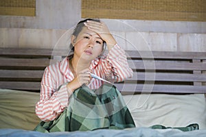 Sweet Asian Korean girl in pajamas covered with blanket sick suffering cold and flu taking temperature with thermometer in bed