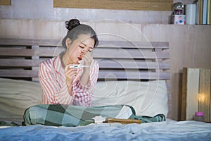 Sweet Asian Korean girl in pajamas covered with blanket sick suffering cold and flu taking temperature with thermometer in bed