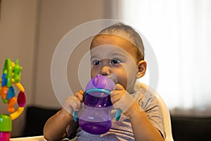 Sweet baby boy sitting in high baby chair and drinking water from baby bottle. Baby at home