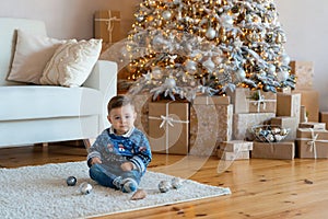 Sweet baby boy sitting on a white wool carpet in decorated living room with a Christmas tree and many gift boxes. First