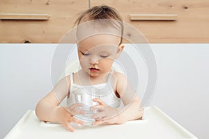 Sweet baby girl holding glass and drinking water. Happy baby sitting in a baby chair in the kitchen