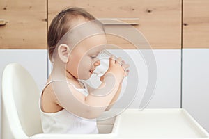 Sweet baby girl holding glass and drinking water. Happy baby sitting in a baby chair in the kitchen