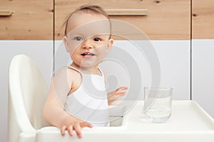 Sweet baby girl holding glass and drinking water. Happy baby sitting in a baby chair in the kitchen