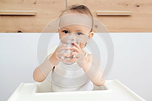Sweet baby girl holding glass and drinking water. Happy baby sitting in a baby chair in the kitchen
