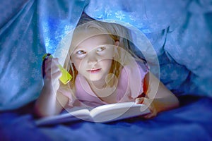 Sweet beautiful and pretty little blond child girl 6 to 8 years old lying under bed covers reading book in the dark with torch fla