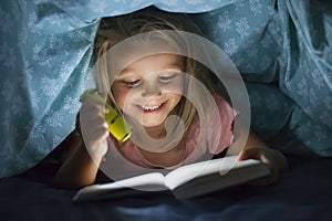 Sweet beautiful and pretty little blond girl 6 to 8 years old under bed covers reading book in the dark at night with torch light