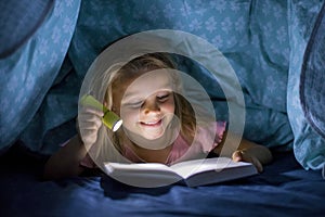 Sweet beautiful and pretty little blond girl 6 to 8 years old under bed covers reading book in the dark at night with torch light