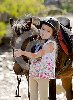 Sweet beautiful young girl 7 or 8 years old hugging head of little pony horse smiling happy wearing safety jockey helmet in summer