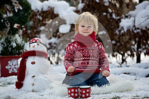 Sweet blond toddler child, boy, playing in garden with snow, making snowman, happy kid winter time