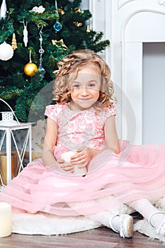 Sweet blonde girl with curls in a pink dress sits in the New Year decorations near a tree decorated with toys