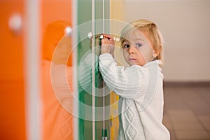 Sweet blonde toddler boy standing in front of a lockers in kindergarden or school hallway