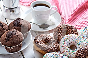 Sweet breakfast with chocolate muffins and donuts, cup of black coffee on white wooden board table. Red dishcloth